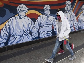 A man wearing a protective face mask walks past a mural of heath care workers painted on a boarded up restaurant in downtown Vancouver.