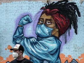 A pedestrian wearing a mask walks past a mural of a woman flexing her muscles while wearing a mask.