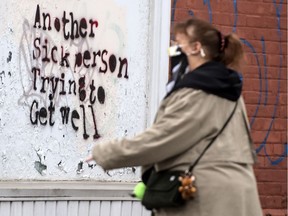 A pedestrian wearing a mask walks past graffiti stating " Another Sick Person Trying To Get Well" in Hamilton during the COVID-19 pandemic.
