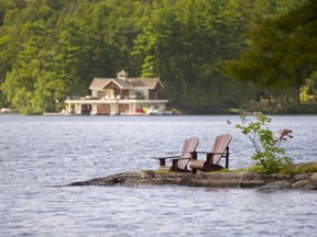 Two Muskoka chairs on a rock formation facing a calm lake. Accross the lake there's a cottage.