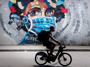File: In Montreal, a man wears a face mask as he cycles by a mural defaced by graffiti objecting to mask usage to protect against the spread of COVID-19