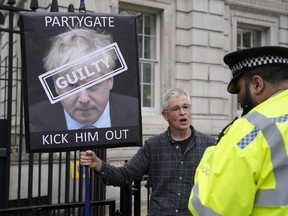 A police officer talks to a protester holding up a sign showing British Prime Minister Boris Johnson, in front of the entrance to Downing Street in London, Wednesday, April 13, 2022. Johnson was fined for breaching COVID-19 regulations following allegations of lockdown parties at government offices. Treasury Chief Rishi Sunak was also fined.