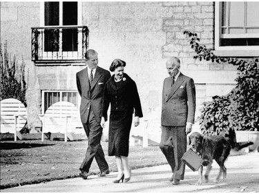 Her Majesty Queen Elizabeth II strolls in the gardens at Rideau Hall, accompanied by His Excellency the Governor General Vincent Massey and H.R.H. Prince Philip. "Duff", a golden retriever, is carrying her handbag.
