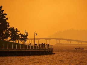 A person travels in a boat past people walking on the boardwalk as smoke from the McDougall Creek wildfire blankets the area on Okanagan Lake, in Kelowna, B.C., Friday, Aug. 18, 2023.
