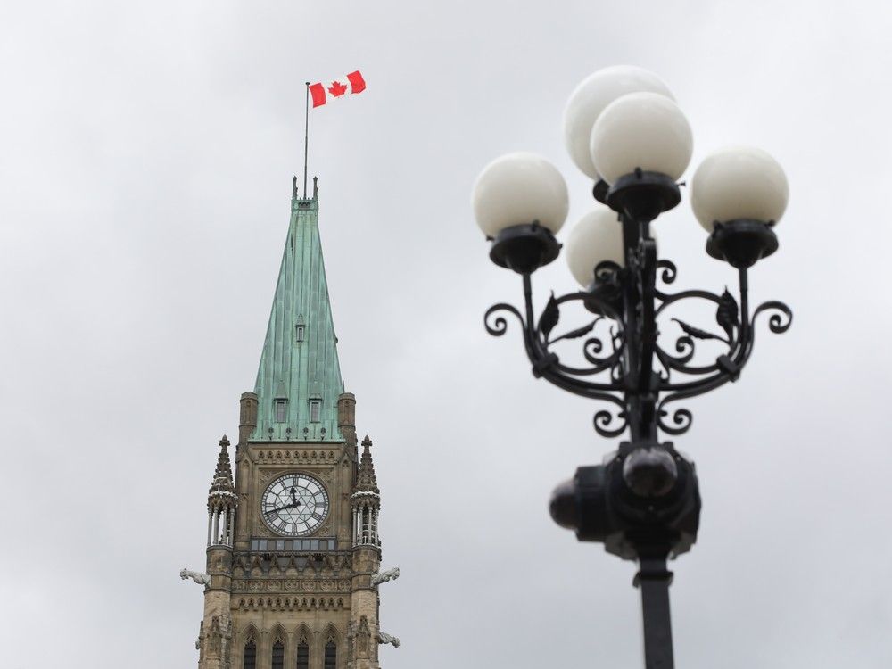 A Canadian flag flies over the Peace Tower clock, with an ornate street lamp in the foreground.
