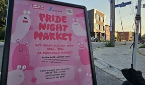 A sandwich board with a pink poster for the Pride Night Market at the corner of a street