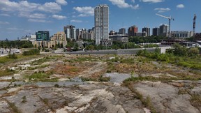 A flat, open stretch of land at LeBreton Flats in downtown Ottawa, bordered by roads and light rail tracks, with the city skyline visible in the background.
