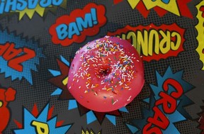 A donut with pink icing and sprinkles on a surface covered in comic book exclamations, like bam! and crunch!
