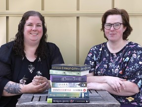 Two friends are sitting side by side behind a pile of books on the table in front of them.