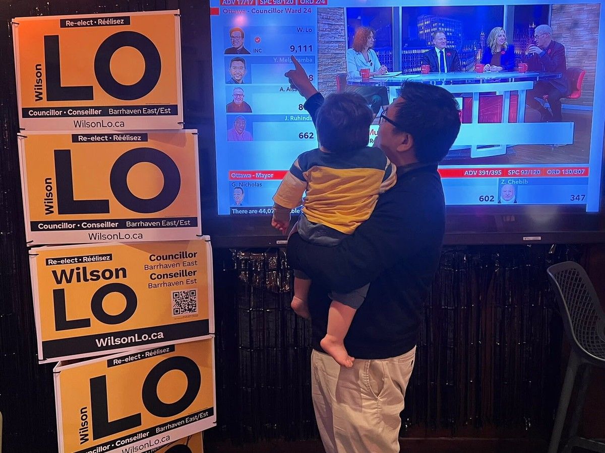 Holding one-year-old son Wesley, Wilson Lo points to his big victory on the TV screen at his re-election party at Heart & Crown's Barrhaven location. Photo by Callum Fraser/Postmedia