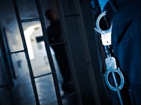 In this stock photo, a guard with handcuffs stand in front of an incarcerated inmate.