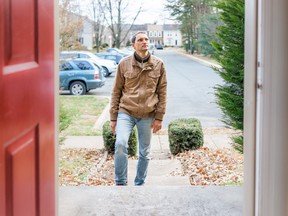 Young man standing on front porch frontyard steps in front of house home buyer looking to buy property real estate customer client in townhouse residential neighborhood, cars parked, door open