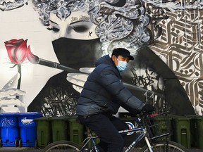A man wearing a protective mask rides his bicycle past a masked mural during the COVID-19 pandemic.