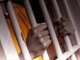 A black man with hands outside the bars of a prison cell.