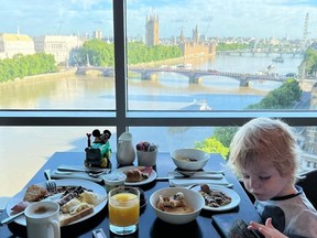 Boy playing on device as he enjoys breakfast in London hotel room overlooking view of the city.