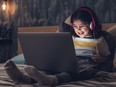 Little girl wearing headphones, sitting on bed in dark bedroom and laughing while looking at laptop.