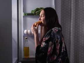 Woman eating an eclair in front of open refrigerator door.