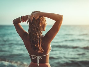 Back of young woman in bikini with raised arms enjoying sunset while looking out at ocean.