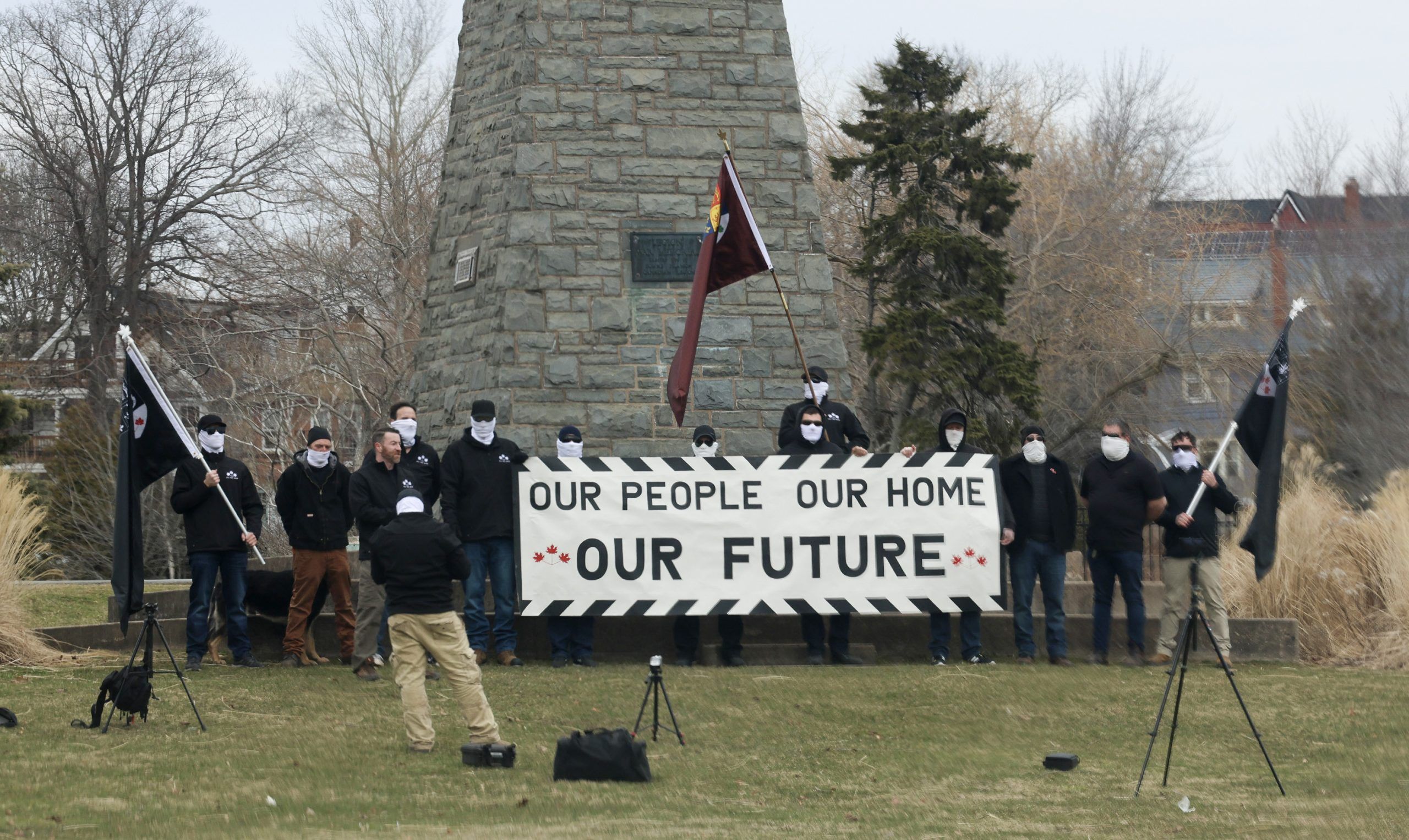 Members of an alt-right group, some with All Thy Sons monogrammed jackets and sporting a version of a modified former Canadian Red Ensign, record a video at the cenotaph at Sullivan's Pond in Dartmouth Sunday.