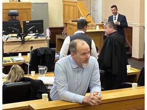 Matthew Kavanagh speaks with a loved one in the gallery during a break in his murder trial at Newfoundland and Labrador Supreme Court in St. John's, Oct. 15 2026. Behind him are his lawyers, Stephen Orr and Iain Hollett, and in the witness box is medical examiner Dr. Chris Hough.