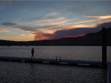 A woman walks on a dock at Kamloops Lake in Savona, B.C., as smoke from a wildfire burning near Ashcroft rises in the distance at sunset on Friday July 7, 2017.