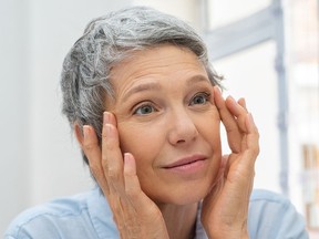 Beautiful senior woman checking her face skin and looking for blemishes. Portrait of mature woman massaging her face while checking wrinkled eyes in the mirror. Wrinkled lady with grey hair checking wrinkles around eyes, aging process.