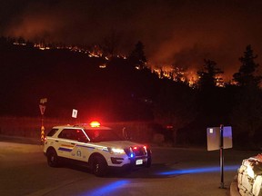 File photo of an RCMP vehicle at an intersection with emergency lights on and wildfire in the background.