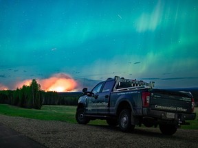 The wildfire forcing thousands to evacuate from a northeast British Columbia town has doubled again in size as the blaze grows merely a few kilometres west of city limits. The Aurora Borealis shines overhead of a B.C. Conservation Officer Service vehicle near the junction of highways 97 and 77, as a wildfire burns in the background near Fort Nelson, B.C., in a Saturday, May 11, 2024, handout photo.