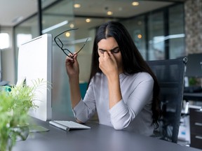 Portrait of businesswoman sitting at computer in office holding glasses and rubbing eyes.