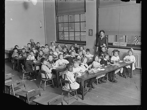 Children sit at long tables in a classroom while their teacher looks on, sometime between 1919 and 1929.
