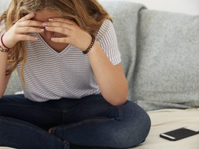 In this stock photo, a depressed teenage girl sits with her head in her hands as her smartphone sits on the couch next to her.
