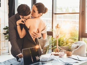 Young man in suit kissing beautiful woman in black dress on shoulder in his office