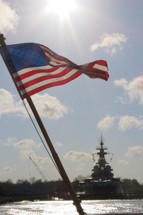Stars and Stripes flag on stern of U.S. Coast Guard ship at Wilmington, N.C. with 1940 battleship USS North Carolina across Cape Fear River, where memorial attracts 300,000-plus visitors a year. (Ian S. Robertson)