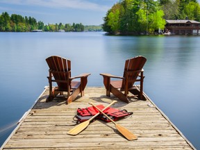 Two Muskoka chairs on a wooden dock facing a lake. Paddles and life jackets are visible on the dock. Across the calm water there is a brown cottage.