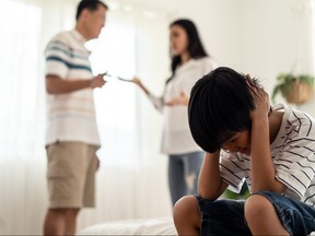 Asian boy kid sitting and crying on bed while parents having fighting or quarrel conflict at home. Child covering ears with hands do not want to hear the violence argument. Domestic problem in family.