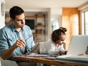 Stay at home father feeling stressed while baby sitting and trying to work while his small daughter is using his laptop.