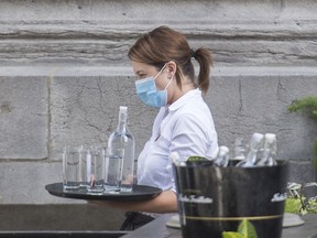 A server wears a face mask as she carries a tray of glasses and water at a restaurant in Montreal, Sunday, July 5, 2020. As the Canadian economy continues to adapts to the reality of the COVID-19 pandemic, some restaurants in the country's most populous city are saying goodbye to a service industry staple: tipping.