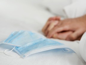 closeup of a pair of surgical masks on a bed set with white bedsheets, where two people are gripping their hands in the background