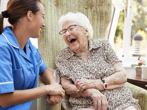 Senior Woman Sitting In Chair And Laughing With Nurse In Retirement Home