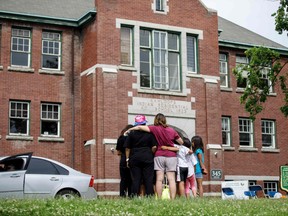 Jennifer Nickel holds her daughters as they look up at the former Kamloops Indian Residential School where flowers and cards have been left as part of a growing makeshift memorial to honour the 215 children whose remains have been discovered buried near the facility, in  Kamloops, B.C., on June 3, 2021.