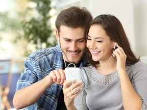 Happy couple sharing music from smart phone sitting on a couch at home.
