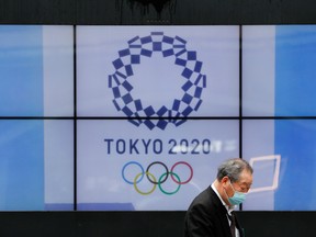 A passerby wearing a protective face mask walks past a screen showing the logo of the 2020 Olympic Games in Tokyo, April 14, 2021.