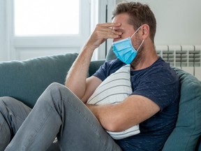 Sad man with protective face mask at home living room couch feeling tired and worried suffering depression amid coronavirus lockdown and social distancing. Mental Health and isolation concept.