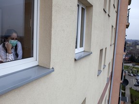 A woman wearing a face mask looks out of a window while quarantining.