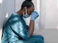 Tired depressed female african scrub nurse wears face mask blue uniform gloves sits on hospital floor. Exhausted sad black doctor feels burnout stress of corona virus frontline protection pray at work