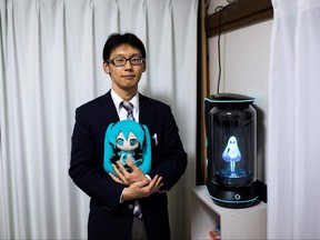 In this photograph taken on November 10, 2018 Japanese Akihiko Kondo poses next to a hologram of Japanese virtual reality singer Hatsune Miku as he holds the doll version of her at his apartment in Tokyo, a week after marrying her. - Akihiko Kondo, who is an administrator at a school, married to a Japanese virtual reality singer called Hatsune Miku in early November 2018. The bride is a computer animation with saucer eyes and lengthy aquamarine pigtails who performs to adoring fans as virtual reality character.