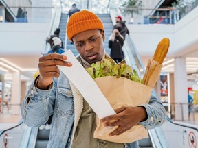 Surprised African-American man in denim jacket looks at receipt total in sales check holding paper bag with products in mall