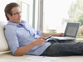 Man reclining on sofa using remote control and laptop