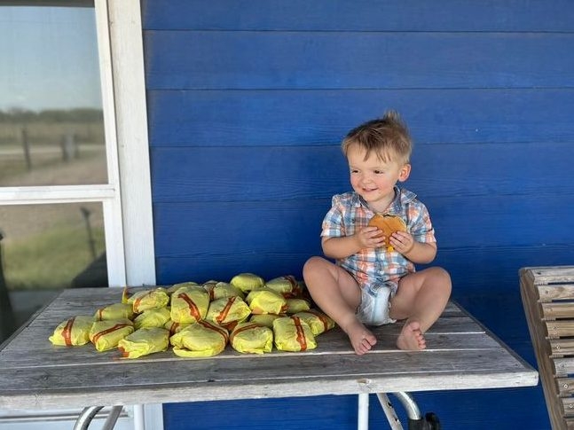 Smiling toddler sitting on table holding cheeseburger with a pile of wrapped cheeseburgers in yellow paper next to him.