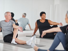 Senior man sitting on mat and doing relaxation exercises during yoga class with other people in studio.